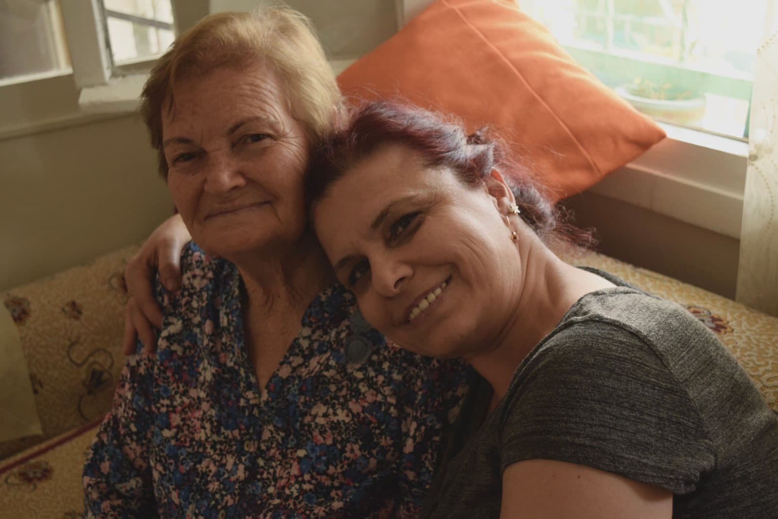 Una mujer sentada junto a su madre en casa, con el brazo sobre sus hombros.